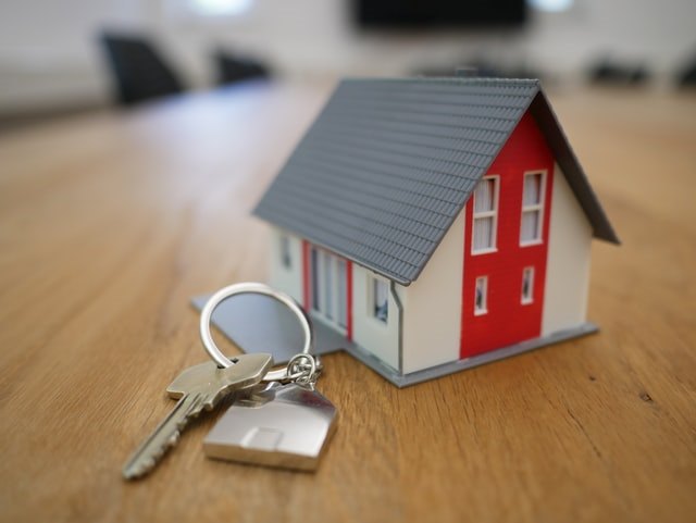 Small model of a red, white and grey house placed next to a set of keys on a brown table.