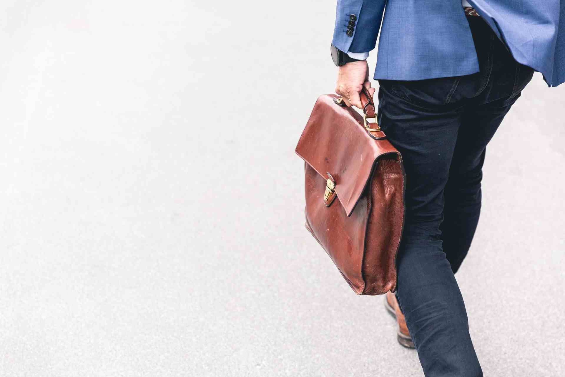 Man wearing blue suit jacket and navy trousers walking away with brown briefcase.