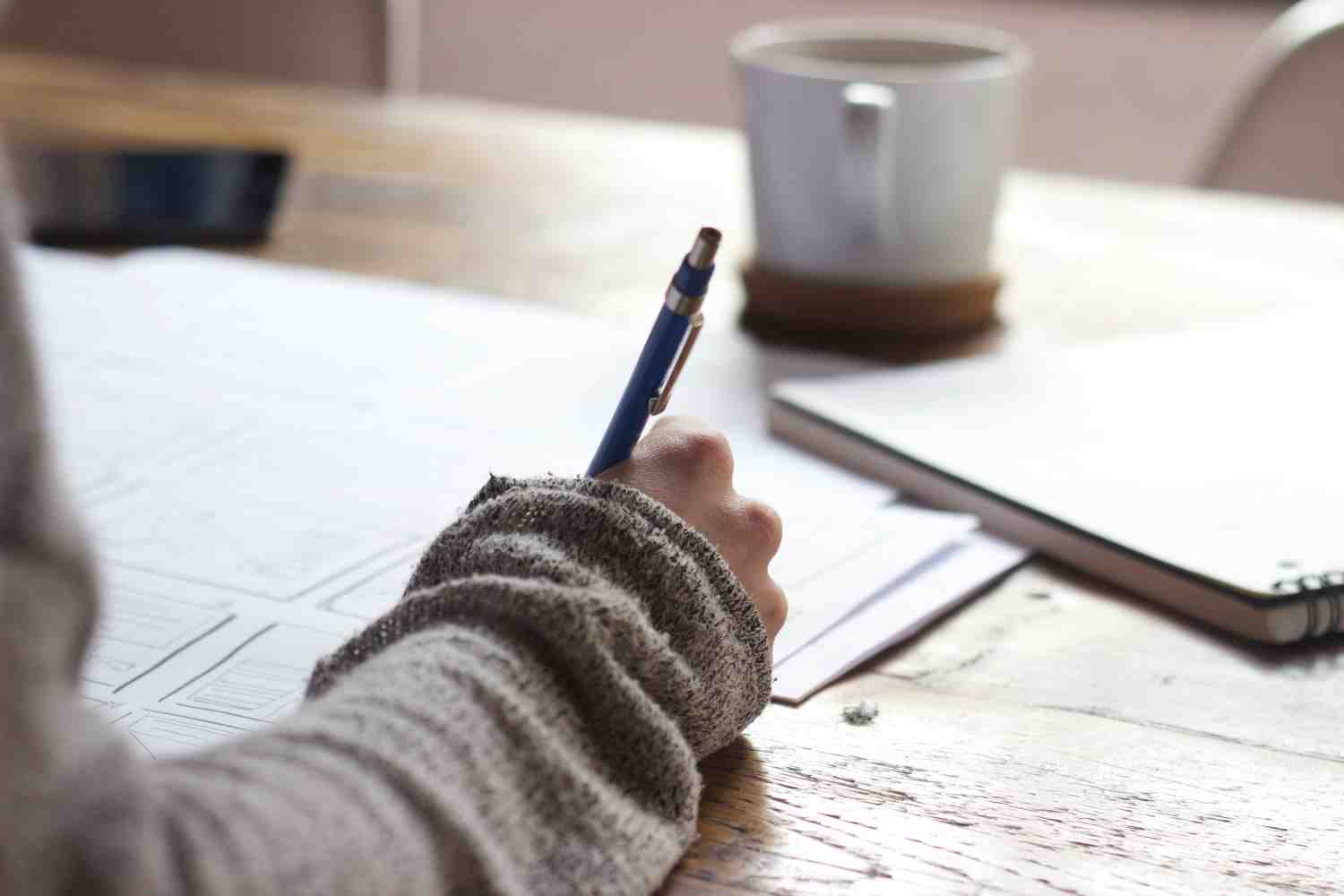 Wooden table with a white mug and contracts on top. A person is signing the documents with a pen.