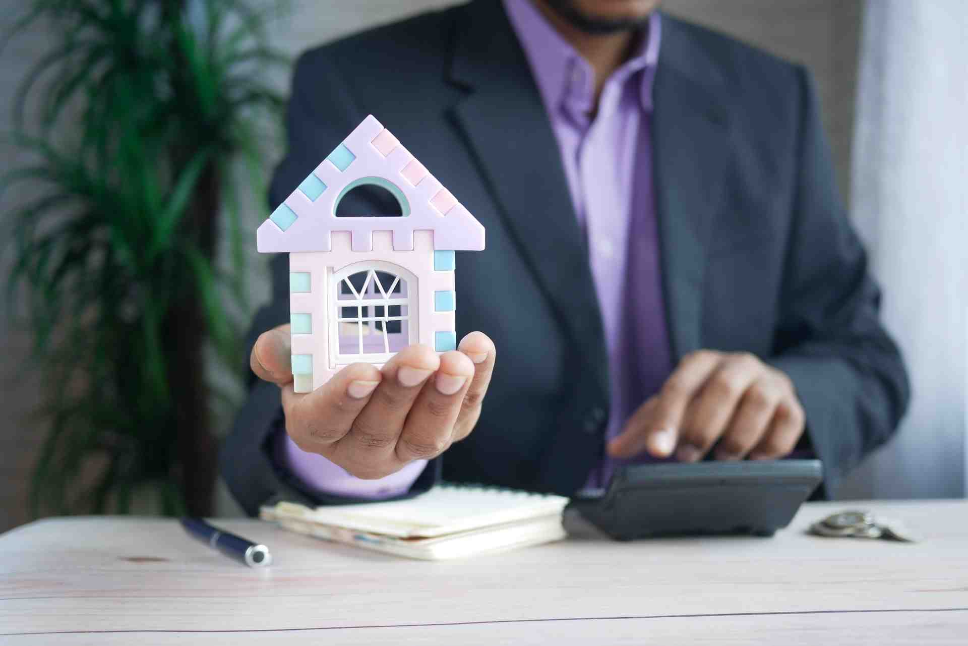 A man in a dark suit and purple shirt is sat at a light wooden desk with a notepad, calculator and pen. He his holding out his hand and holding a small model house in it.