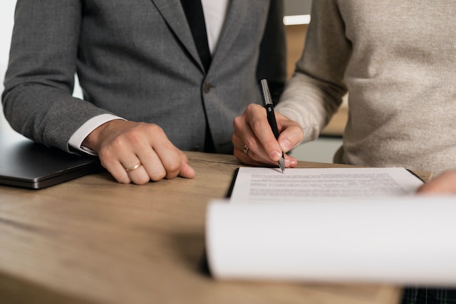 Man in grey suit standing next to man in grey jumper signing papers.